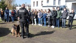 Na zewnątrz komendy umundurowany policjant, przewodnik psa służbowego opowiada młodzieży szkolnej o specyfice swojej pracy. Na zdjęciu widoczny też jest pies służbowy.