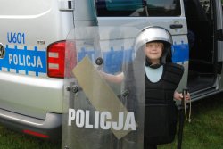 chłopiec ubrany jest w sprzęt policjanta biorącego udział w zabezpieczeniu imprez masowych, za nim znajduję się policyjny oznakowany radiowóz