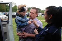 Umundurowana policjantka wręcza dziecku element odblaskowy, dziecko jest na rękach mężczyzny.
