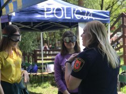 Strażniczka Miejska rozmawia z dwoma nastoletnimi dziewczynami, nastolatki mają założone alkogogle, w tle widoczny jest niebieski namiot z napisem POLICJA.