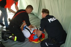 Umundurowany policjant wraz ze strażakiem udzielają pomocy osobie leżącej na desce ortopedycznej, wszyscy znajduję się w namiocie