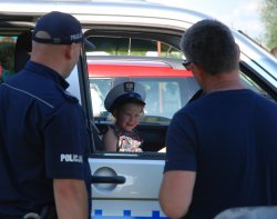 dziewczynka w białej policyjnej czapce będąc w oznakowanym radiowozie pozuje do zdjęcia, po lewej stronie tyłem do zdjęcia stoi umundurowany policjant, po prawej mężczyzna