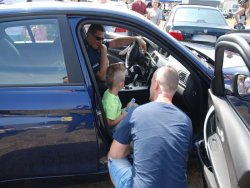 Policjanci drogówki prezentują radiowóz na zlocie miłośników BMW. W środku radiowozu siedzi policjant i chłopiec, przed radiowozem przykucnął jego tata.