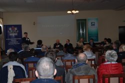 Umundurowany policjant omawia zagrożenia związane z działaniami oszustów. Na zdjęciu widoczni są też uczestnicy debaty.